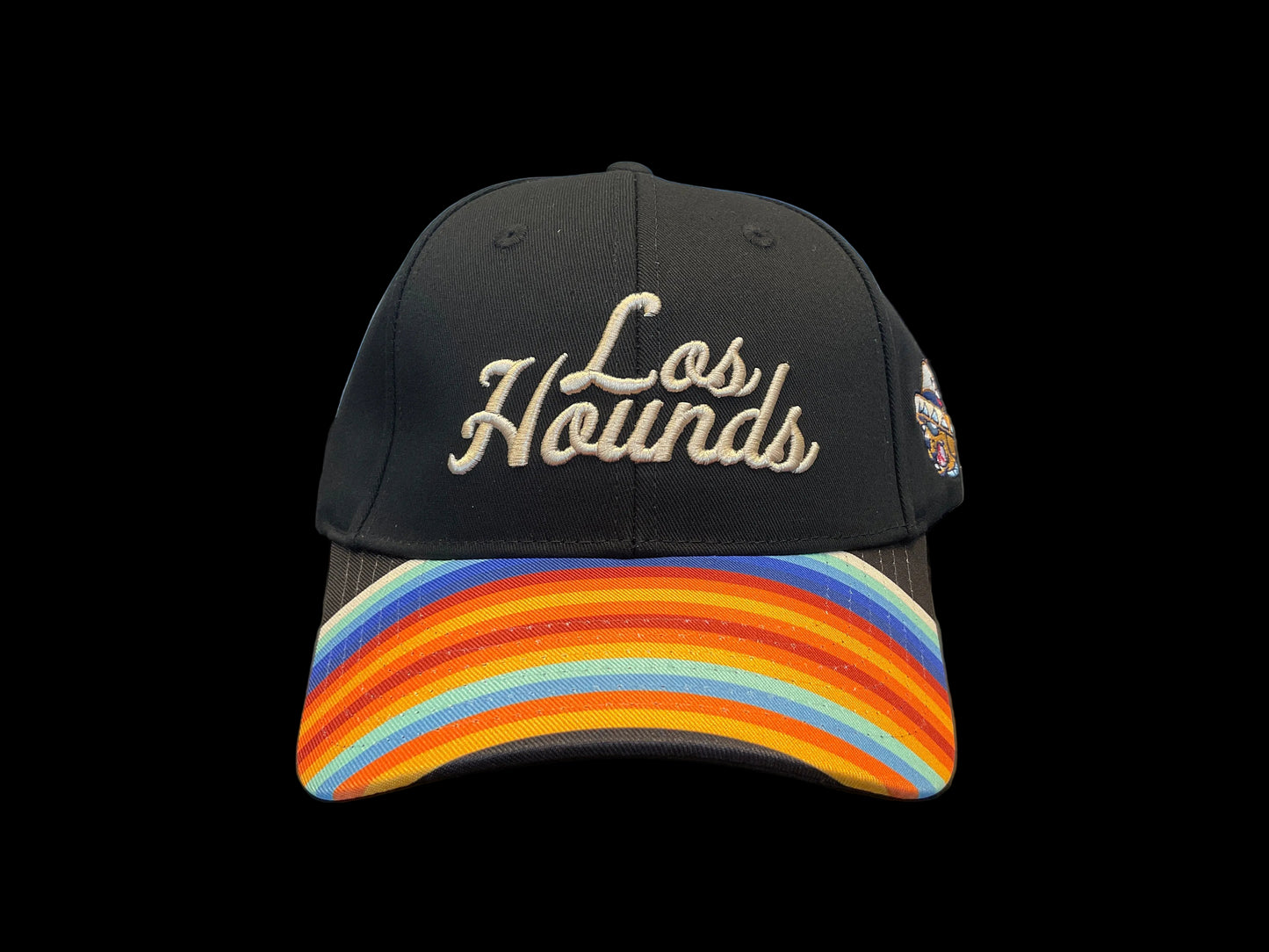 Black cap with 'Los Hounds' text and colorful brim on a table with shirts in the background